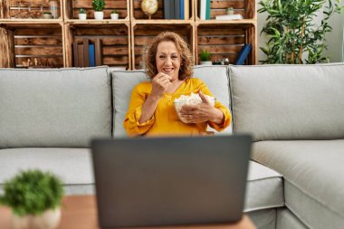 Orta yaşlı, beyaz bir kadın film seyrediyor. Evdeki kanepede oturuyor..