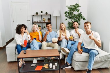 Group of people eating snack sitting on the sofa at home smiling happy and positive, thumb up doing excellent and approval sign 