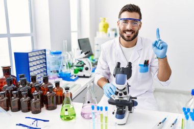 Handsome hispanic man working at scientist laboratory smiling with an idea or question pointing finger with happy face, number one 