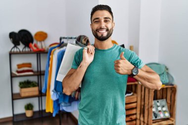 Satış mağazasında sakallı, alışveriş torbası tutan genç yakışıklı bir adam mutlu bir el hareketi yapıyor. Kameraya bakıp başarıyı gösteren ifade. 