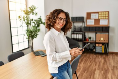 Orta yaşlı İspanyol kadın, ofiste dokunmatik ped kullanarak çalışırken kendine güvenerek gülümsüyor.