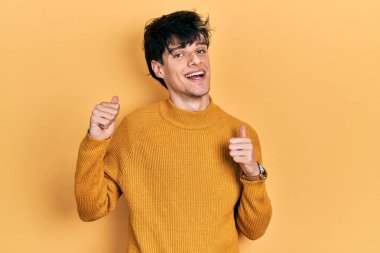 Yakışıklı hippi genç adam gündelik sarı süveter giyiyor. Arkayı işaret ediyor. El ve baş parmakları havada, kendine güvenen. 