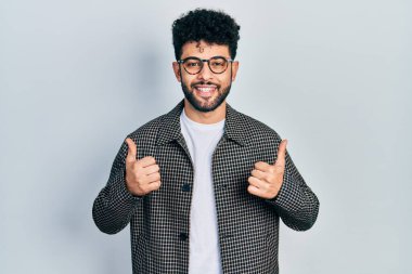 Young arab man with beard wearing glasses success sign doing positive gesture with hand, thumbs up smiling and happy. cheerful expression and winner gesture. 