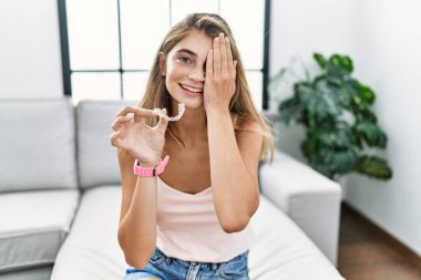 Young blonde woman holding invisible aligner orthodontic covering one eye with hand, confident smile on face and surprise emotion. 