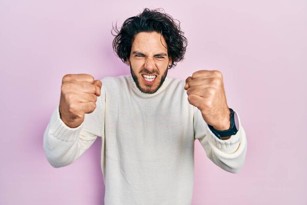Handsome hispanic man wearing casual white sweater angry and mad raising fists frustrated and furious while shouting with anger. rage and aggressive concept. 