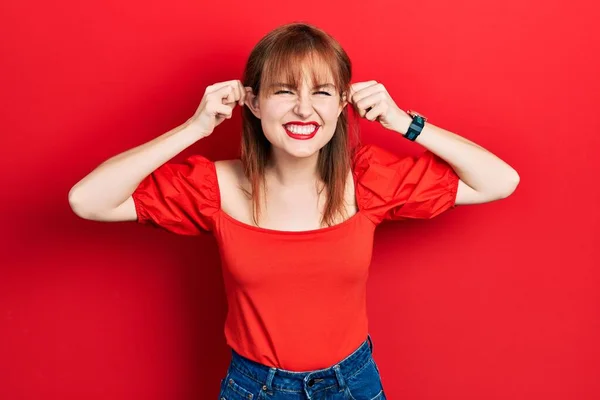 Kırmızı Tişörtlü Kızıl Saçlı Genç Kadın Gülümsüyor Kulakları Parmaklarıyla Çekiyor — Stok fotoğraf