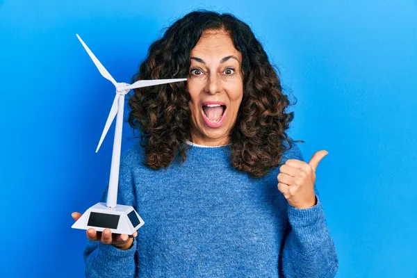 再生可能エネルギーのためのソーラー風車を保持している中年のヒスパニック女性口を開けて幸せな笑顔側に親指を指す — ストック写真