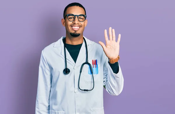 身穿医生制服和听诊器的年轻的非洲裔美国人 带着自信和快乐的笑容 用五号手指显示和指尖 — 图库照片