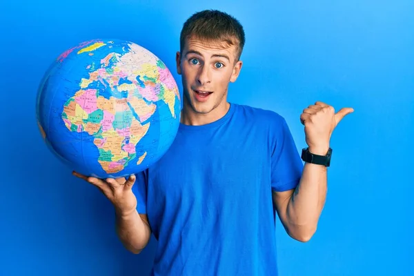 Young Caucasian Man Holding World Ball Pointing Thumb Side Smiling — Stockfoto