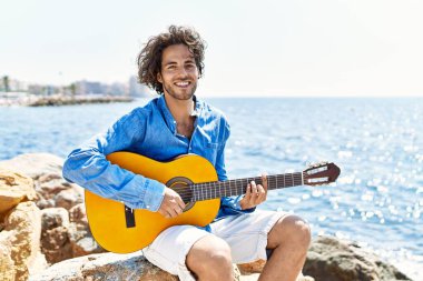 Genç İspanyol adam klasik gitar çalıyor plajda rock 'ın üzerinde oturuyor..