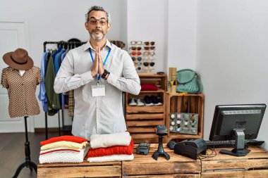 Middle age hispanic man working as manager at retail boutique praying with hands together asking for forgiveness smiling confident. 