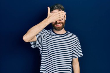 Sakallı, çizgili tişörtlü, gözlüklü, gülümseyen ve elleri yüzünde, şaşırtmak için gözlerini kapatan beyaz bir adam. kör kavram. 