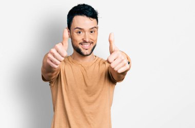 Sakallı, sıradan tişörtlü İspanyol bir adam elinle olumlu jest yapmayı onaylıyor, gülümsüyor ve başarı için seviniyor. kazanan jest. 