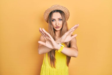 Young blonde girl wearing summer hat rejection expression crossing arms doing negative sign, angry face 
