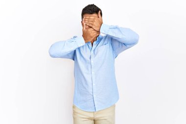 Young hispanic man wearing business shirt standing over isolated background covering eyes and mouth with hands, surprised and shocked. hiding emotion 