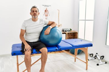 Middle age hispanic man at pain recovery clinic holding pilates ball looking at the camera blowing a kiss on air being lovely and sexy. love expression. 