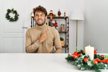 Noel süslemeleri ile masada oturan sakallı yakışıklı genç adam endişeli ve endişeli bir yüz ifadesine işaret ediyor. 
