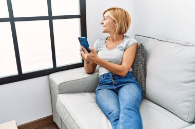 Orta yaşlı sarışın kadın evde akıllı telefon kullanarak gülümsüyor.