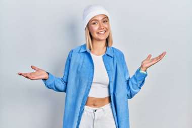 Güzel sarışın kadın yün şapka takıyor gülümsüyor iki elini de açık avuçlarında gösteriyor, sunuyor ve reklam karşılaştırması yapıyor ve dengeyi sağlıyor. 