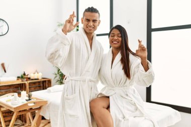 Young latin couple wearing towel standing at beauty center gesturing finger crossed smiling with hope and eyes closed. luck and superstitious concept. 