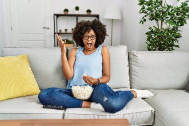 Afro saçlı Afro-Amerikalı kadın evinde patlamış mısır yiyor zaferi mutlu bir gülümsemeyle ve ellerini kaldırarak kazanan ifadeyle kutluyor. 