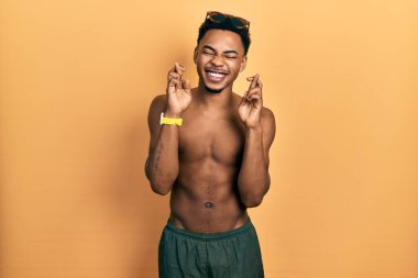 Young african american man wearing swimwear and swimmer glasses gesturing finger crossed smiling with hope and eyes closed. luck and superstitious concept. 