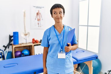 Kısa saçlı, İspanyol bir kadın. Ağrı kesici kliniğinde çalışıyor. Gülümsüyor ve başka tarafa bakıyor.. 