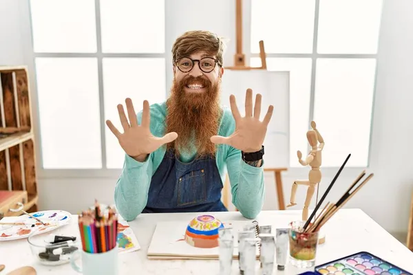 红头发男人 留着长胡子 在艺术工作室里画着黏土碗 用十号手指指指着 面带微笑 自信而快乐 — 图库照片