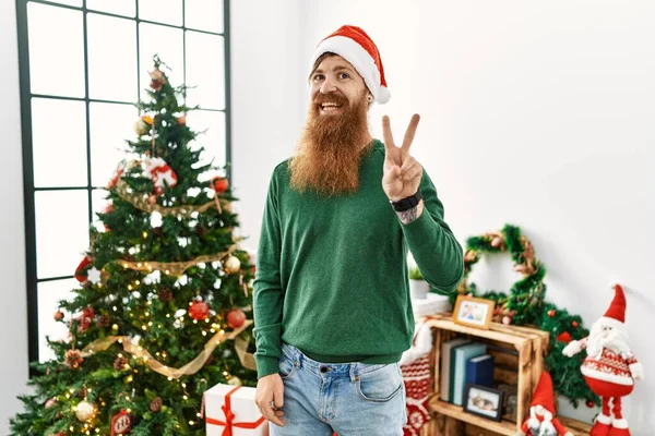 红头发男人留着长胡子 头戴圣诞帽 站在圣诞树旁 用手指指了指二号 面带微笑 自信而快乐 — 图库照片