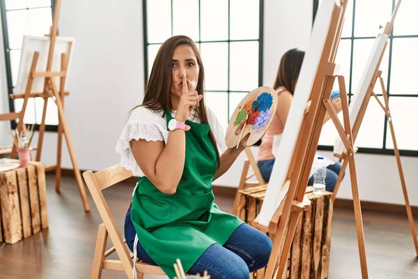 若いヒスパニック系のアーティストの女性がアートスタジオでキャンバスに絵を描く唇に指で静かにするように求めています 沈黙と秘密の概念 — ストック写真
