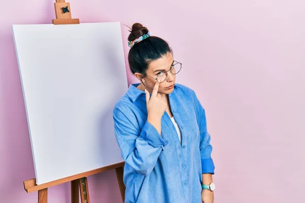 画家のイーゼルのそばに立つ若いヒスパニック系の女性は あなたのジェスチャーを見て目を指して立つ 疑わしい表現 — ストック写真