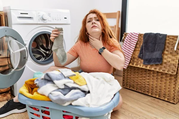 若い赤毛の女性は 痛い首に触れる洗濯機に汚れた洗濯機を入れて インフルエンザ 雲や感染症のための喉の痛み — ストック写真
