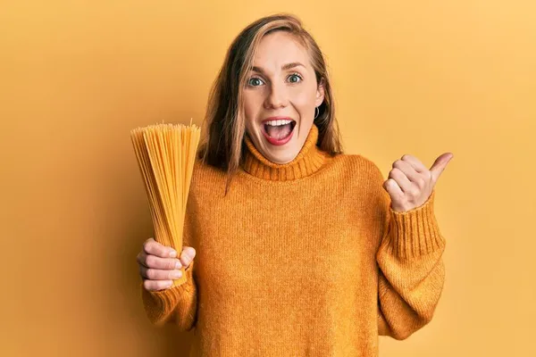 Młoda Blondynka Trzyma Suche Spaghetti Wskazując Kciuk Górę Boku Uśmiechając — Zdjęcie stockowe