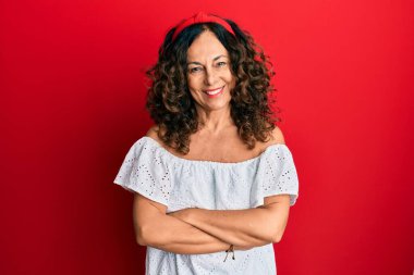 Orta yaşlı İspanyol kadın gündelik kıyafetler giyip kollarını kavuşturup kameraya bakarken gülümsüyor. pozitif kişi. 