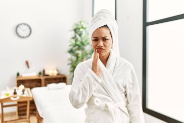 Havlu ve bornoz giyen genç esmer kadın diş ağrısı ya da diş ağrısı yüzünden güzellik merkezinde durup ağız ve acı dolu bir ifadeyle dokunuyor. dişçi 