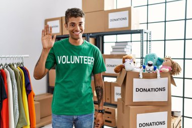 Young handsome hispanic man wearing volunteer t shirt at donations stand waiving saying hello happy and smiling, friendly welcome gesture 