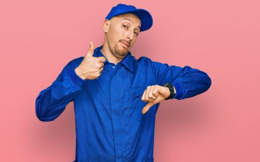 Bald man with beard wearing builder jumpsuit uniform doing thumbs up and down, disagreement and agreement expression. crazy conflict 