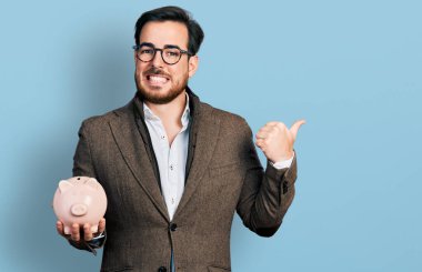 Young hispanic man holding piggy bank pointing thumb up to the side smiling happy with open mouth 