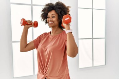 Genç Afrikalı Amerikalı kadın gülümsüyor spor merkezinde halterleri kullanarak kendine güven eğitimi alıyor