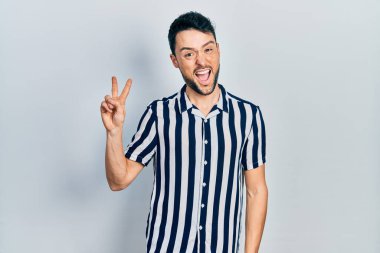 Gündelik kıyafetler giyen, gülümseyen ve kameraya göz kırpan genç İspanyol adam zafer işareti yapıyor. İki numara.. 