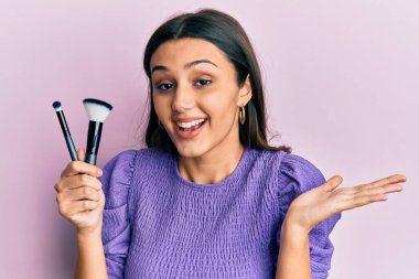 Makyaj fırçası tutan genç İspanyol kadın başarıyı mutlu bir gülümsemeyle kutluyor ve kazanan el kaldırarak ifade ediyor. 