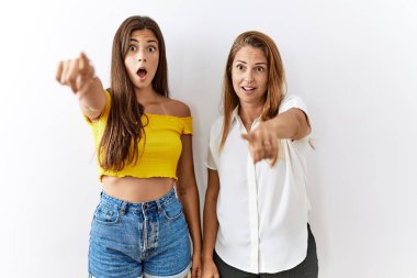 Mother and daughter together standing together over isolated background pointing with finger surprised ahead, open mouth amazed expression, something on the front 