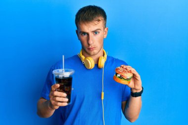 Young caucasian man eating a tasty classic burger and soda depressed and worry for distress, crying angry and afraid. sad expression. 