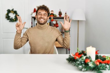 Young handsome man with beard sitting on the table by christmas decoration showing and pointing up with fingers number eight while smiling confident and happy. 