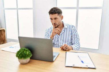 Young handsome man with beard working at the office using computer laptop asking to be quiet with finger on lips. silence and secret concept. 