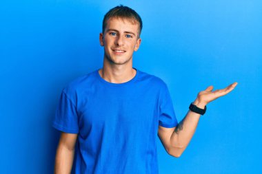 Young caucasian man wearing casual blue t shirt smiling cheerful presenting and pointing with palm of hand looking at the camera. 