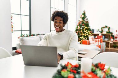 Genç Afro-Amerikan adam dizüstü bilgisayar kullanıyor Noel ağacının yanında oturmuş komik bir ifadeyle dilini dışarı çıkarıyor. duygu kavramı. 