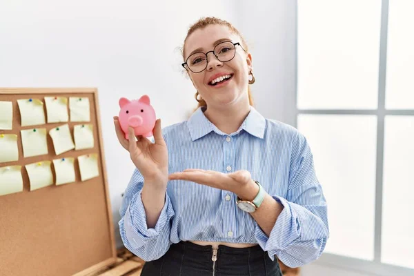 Young Redhead Woman Holding Piggy Bank Office Smiling Happy Pointing — Fotografia de Stock