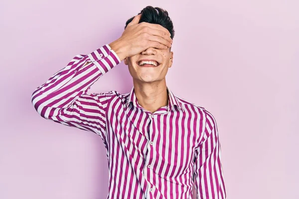 カジュアルな服を着た若いヒスパニック系の男が笑顔で 驚きのために目をカバー顔に手で笑っています 視覚障害の概念 — ストック写真