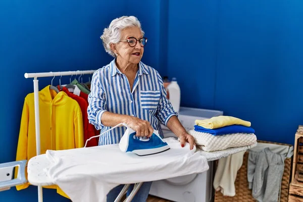 Senior grey-haired woman smiling confident ironing clothes at laundry room